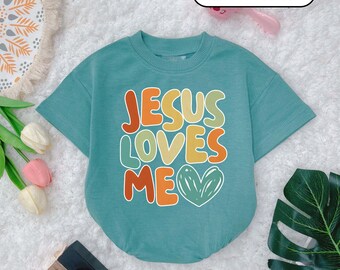 May include: A cadet blue t-shirt with the text "JESUS LOVES ME" in colorful, retro-style lettering. The shirt has short sleeves and a round neckline.
