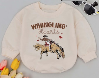 Op de afbeelding: Crèmekleurige babyromper met lange mouwen. De voorkant heeft een afbeelding van een cowboy op een paard, met de woorden "WRANGLING Hearts" in bruin. Rode harten zijn verspreid rond het ontwerp. Het woord "CREAM" staat in een witte ovaal bovenaan.