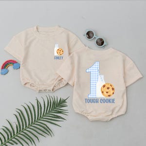 May include: Two beige baby rompers with short sleeves. One has "FINLEY" printed below a milk bottle and cookie graphic. The other has a blue gingham "1" and the words "TOUGH COOKIE" below a milk bottle and cookie graphic.