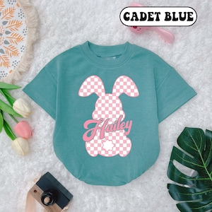 May include: A teal blue short-sleeved t-shirt with a pink checkered bunny graphic and the name "Hailey" in a pink retro font. The shirt is on a white fuzzy surface.