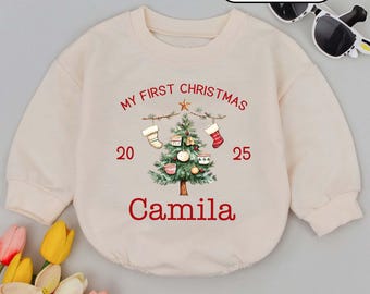 May include: Cream-colored baby romper with long sleeves. The front features a Christmas tree design with the text "MY FIRST CHRISTMAS" above it, the year "2025" and the name "Camila" below. The romper has a round neckline.