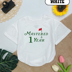 May include: White short-sleeved t-shirt with green text that says "MASTERED 1 YEAR" and a red flag graphic.