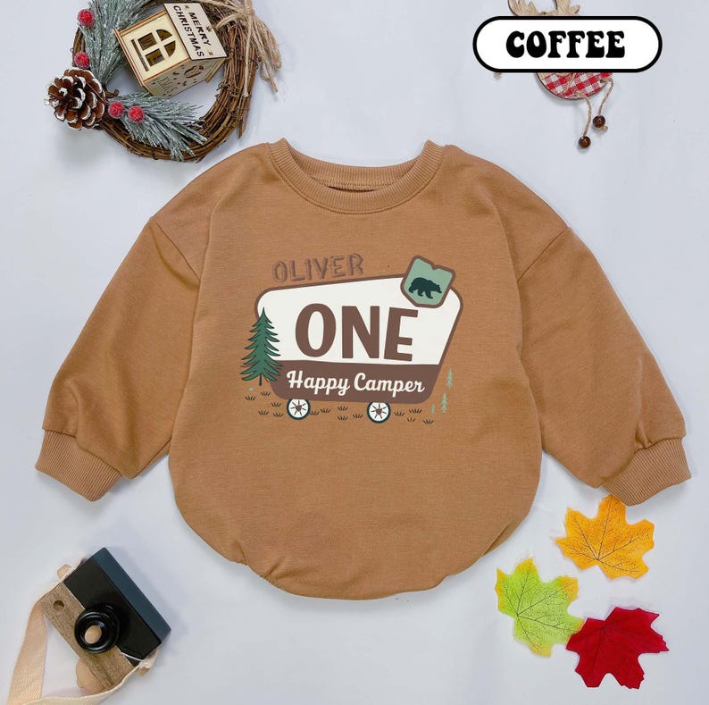 May include: A brown long-sleeved baby bodysuit with the text "OLIVER ONE Happy Camper" and a graphic of a bear, a tree, and a camper. The word "COFFEE" is in a white oval. The bodysuit is surrounded by fall leaves and a Christmas wreath.