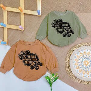 May include: Two baby rompers, one in olive green and the other in tan, each featuring a black graphic of a monster truck and the text "FUTURE MONSTER TRUCK DRIVER". The rompers are made of soft material.