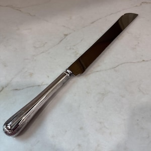 May include: A silver-toned serrated bread knife featuring a long, polished metal blade and a rounded, fluted handle with decorative detailing at the end. The knife has a classic design with a straight bolster connecting the handle to the blade.