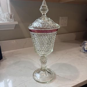 Puede incluir: Un cuenco de cristal transparente con tapa extraíble y base de pedestal, que presenta una textura de patrón de diamantes en el cuerpo y la tapa. La pieza incluye una banda horizontal distintiva de color rosa oscuro cerca del borde.