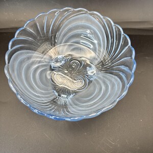 May include: A clear glass serving dish with a light blue tint. The dish has a central, raised section and a scalloped edge. The design features a swirling pattern radiating from the center, creating a decorative and elegant look.