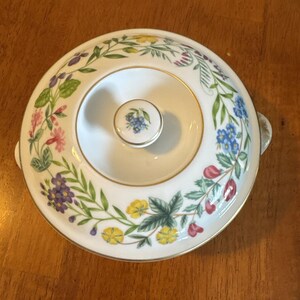 May include: A white ceramic serving dish with a lid, adorned with a floral pattern in shades of purple, pink, yellow, and blue. The dish has a gold rim and a small knob on the lid. The handles are small and curved.
