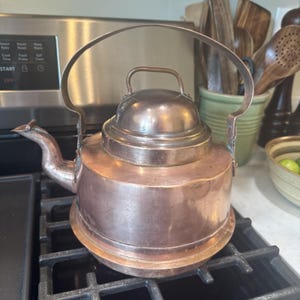May include: A vintage copper kettle with a large arched handle and a small handle on the lid. The kettle is sitting on a black stovetop. The copper has a warm, reddish-brown hue, suggesting age and use. The spout is pointed and the kettle has a rounded base.