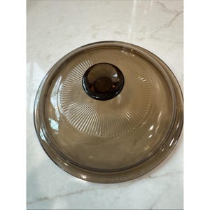 May include: A round, transparent brown glass lid featuring a central knob handle and a radiating sunburst pattern of fine, etched lines extending from the centre towards the outer rim. The lid has a smooth, curved surface and a raised edge.