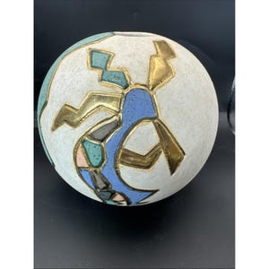 May include: A spherical, off-white ceramic vase with a textured surface. The vase features a geometric design in gold, blue, green, and pink. The design is abstract and covers a large portion of the vase's surface. The vase has a small opening at the top.