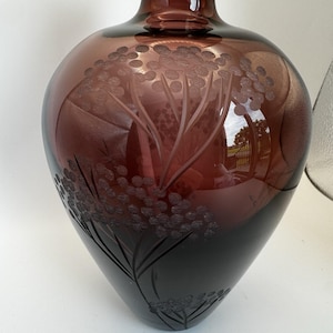 May include: A large, dark purple glass vase with a rounded body and a narrow neck. The vase features an etched floral design, with detailed stems and clusters of small, round shapes. The glass has a gradient effect, transitioning from a lighter purple at the top to a darker shade at the base.