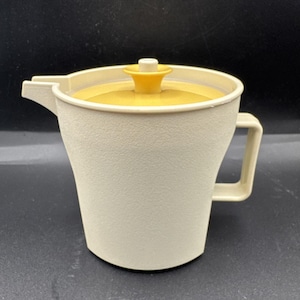 May include: A vintage cream-colored pitcher with a yellow lid and handle. The pitcher has a textured surface and a spout for pouring. The lid is topped with a yellow knob. The handle is a rectangular shape.