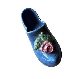Pode incluir: Um vaso azul em forma de sapato de cerâmica com uma rosa rosa e folhas verdes. O vaso tem um acabamento salpicado e um interior oco. O adorno floral decorativo está na lateral do sapato.