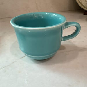 Può includere: Una tazza in ceramica color turchese tinta unita con una finitura liscia e lucida, caratterizzata da una forma a ciotola ampia e arrotondata, un bordo svasato e un manico curvo e robusto fissato lateralmente, dal colore blu-verde uniforme.