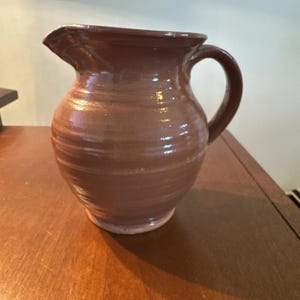 May include: A brown ceramic pitcher with a handle and a spout. The pitcher has a rounded body and a smooth, glossy finish. The pitcher is sitting on a wooden surface.