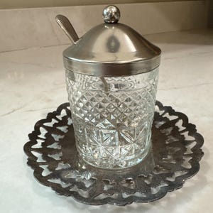 May include: A clear glass jar with a diamond-cut pattern, featuring a silver-toned metal lid with a small knob and a matching silver-toned spoon. The jar rests on a circular silver-toned metal plate with an intricate, openwork scalloped edge.