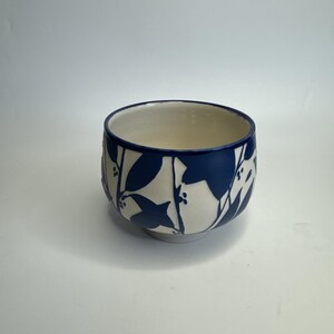 May include: A ceramic bowl with a white interior and a navy blue exterior. The bowl is decorated with a repeating pattern of white leaves and berries against the navy blue background. The bowl is round and has a small base.