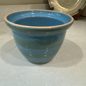 May include: A ceramic planter with a glossy finish featuring horizontal bands of light blue and muted teal. The pot has a wide, flared rim with an exposed terracotta-colored edge, a tapered body, and a smooth, rounded interior surface.