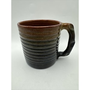 Puede incluir: Una taza de cerámica hecha a mano con un diseño acanalado. La taza hace la transición de un borde marrón oscuro a una base negra, con tonos de verde y marrón intermedios. El asa es negra y tiene una forma única.