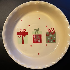 May include: A cream-colored pie plate with a scalloped edge. The plate is decorated with three gift boxes in red and green, and red polka dots. A festive design for holiday baking.