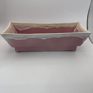 May include: A rectangular planter with a pink body and a textured surface. The rim is a light beige color with a white edge. The planter has a low profile and is elevated by small feet.