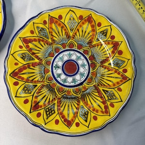 May include: A decorative ceramic plate with a vibrant yellow background and a detailed floral design. The plate features a scalloped edge and a central medallion with red, blue, and white accents. The plate's diameter is approximately 10 inches.
