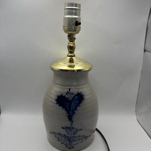 May include: A stoneware lamp with a blue heart design. The lamp has a brass base and a neutral-toned ceramic body. The heart is hand-painted, and the lamp has a standard light socket.