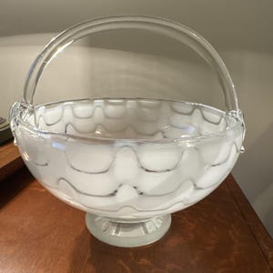 May include: A clear glass basket with a handle, featuring a white interior with a wavy, gray pattern. The basket sits on a round base and is displayed on a wooden surface. The handle is also clear glass.
