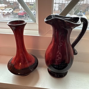 May include: Two ceramic vases with a glossy finish. One is a tall, slender vase with a rounded base. The other is a pitcher with a handle. Both are shades of red and brown, with a dark base.