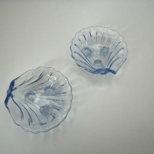 May include: Two light blue glass seashell-shaped dishes. Each dish has a scalloped edge and three small feet. The interior features a textured design. These decorative dishes are ideal for serving small portions or as decorative accents.