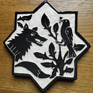 Op de afbeelding: Een keramische tegel met een zwart-wit ontwerp. De tegel heeft de vorm van een achtpuntige ster. Het ontwerp toont een wolf en een vogel met een tak met bladeren. De tegel heeft een zwarte rand.