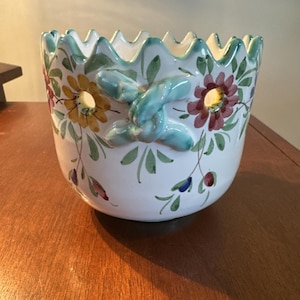 May include: A white ceramic planter with a scalloped rim and floral design. The planter features yellow, red, and blue flowers with green leaves. Two turquoise-colored handles are on the side.
