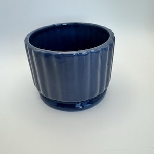 May include: A dark blue ceramic planter with a ribbed design. The planter has a wide base and a slightly flared top.