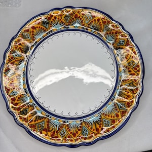 May include: A decorative ceramic plate with a scalloped edge. The plate features a white center, a blue border, and a detailed pattern in yellow, red, and turquoise. The design includes geometric and floral elements.