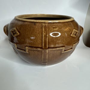 Könnte beinhalten: Eine braune Keramikschale mit glänzendem Finish. Die Schale hat zwei Griffe und geometrische Muster. Die Außenseite weist Rauten- und Rechteckformen auf. Der Innenraum ist dunkler braun.