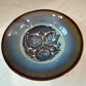 Może przedstawiać: Okrągła ceramiczna miska z reaktywnym szkliwem, posiadająca ciemnobrązowy brzeg, który przechodzi w odcienie jasnego błękitu i szarości. Środek miski zdobi ciemny, teksturowany wzór z trzema płatami, wypukłymi krawędziami i błyszczącymi akcentami.