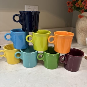 Op de afbeelding: Een stapel van acht keramische mokken in verschillende kleuren: marineblauw, lichtblauw, geel, limoengroen, oranje, blauwgroen, groen en bordeauxrood. Elke mok heeft een afgerond oor en een licht uitlopende rand. De mokken zijn in een piramidevorm gerangschikt.