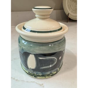 May include: A ceramic canister with a lid, featuring a cream-colored base and a dark green hand-painted band around the lid. The body of the canister displays a stylized, abstract dark charcoal and sage green brushstroke pattern on a cream background.