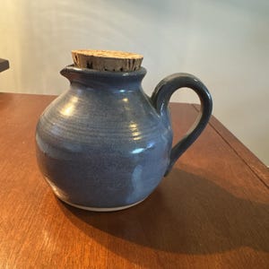 May include: A blue ceramic pitcher with a handle and a cork stopper. The pitcher has a round body and a spout. The cork stopper is made of natural cork. The pitcher is sitting on a wooden surface.