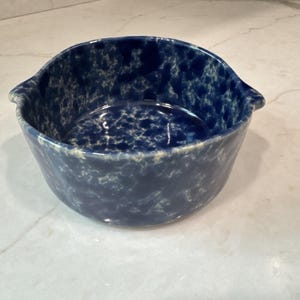 May include: A round ceramic bowl featuring a deep blue and white mottled sponge-painted pattern. The bowl has a smooth, glossy finish and includes two small, integrated handles on opposite sides of the rim, providing a secure grip for handling.