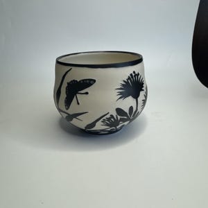 May include: A ceramic bowl with a cream-colored exterior and a black rim. The bowl is decorated with black silhouettes of a butterfly, flowers, and foliage. The design wraps around the exterior of the bowl.