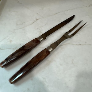 May include: A carving knife and a two-pronged carving fork with metallic silver blades and brown marbled resin handles. The utensils feature a smooth, polished finish on the handles and a sharp, pointed design for the knife and fork tines.