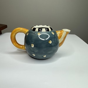 May include: A ceramic teapot with a blue body adorned with white polka dots. The handle is yellow, and the spout is orange and white striped. The top rim features a black and white checkered pattern. A unique and decorative kitchenware item.
