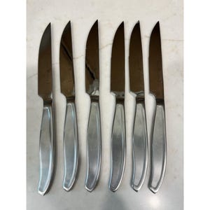 May include: A set of six stainless steel steak knives arranged in a vertical row. Each knife features a pointed, sharp blade and a smooth, polished metal handle with a slightly curved design. The knives have a uniform, reflective silver finish.