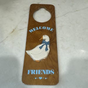 Pode incluir: Um cabide de porta de madeira castanha com uma figura de ganso branco em relevo com um laço azul. O texto WELCOME está impresso em azul claro acima do ganso, e a palavra FRIENDS está impressa em azul claro abaixo, com três corações.