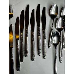 May include: A collection of stainless steel cutlery, comprising knives, spoons, and a fork, displayed on a white surface. The knives feature a decorative handle design. The spoons have a rounded bowl.