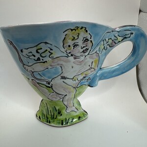 May include: A hand-painted ceramic teacup with a light blue exterior and a green base. The cup features a cherub design with a bow and arrow. The handle and rim are also light blue. The cherub is depicted in a whimsical style.