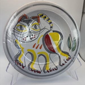 May include: A ceramic plate with a hand-painted cat design. The cat is depicted in a whimsical style with yellow, red, and black accents on a gray background. The plate has a gray rim and is displayed on a clear stand.