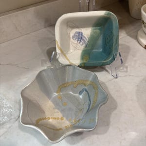 May include: Two ceramic bowls with speckled off-white glaze. The top bowl is square with rounded corners, featuring a teal side accent and blue markings. The bottom bowl has a scalloped, flower-like rim with light blue and yellow decorative swirls.